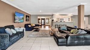 A spacious living room with a large TV, multiple sofas, and a kitchen area in the background. - Spacious Villa with Heated Pool, Trampoline, BBQ, Tennis, Air Hockey (Hollywood)