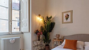 Ein Schlafzimmer mit Bett, Heizkörper, Fenster, Wandleuchte, gerahmtem Bild und einem kleinen Beistelltisch.