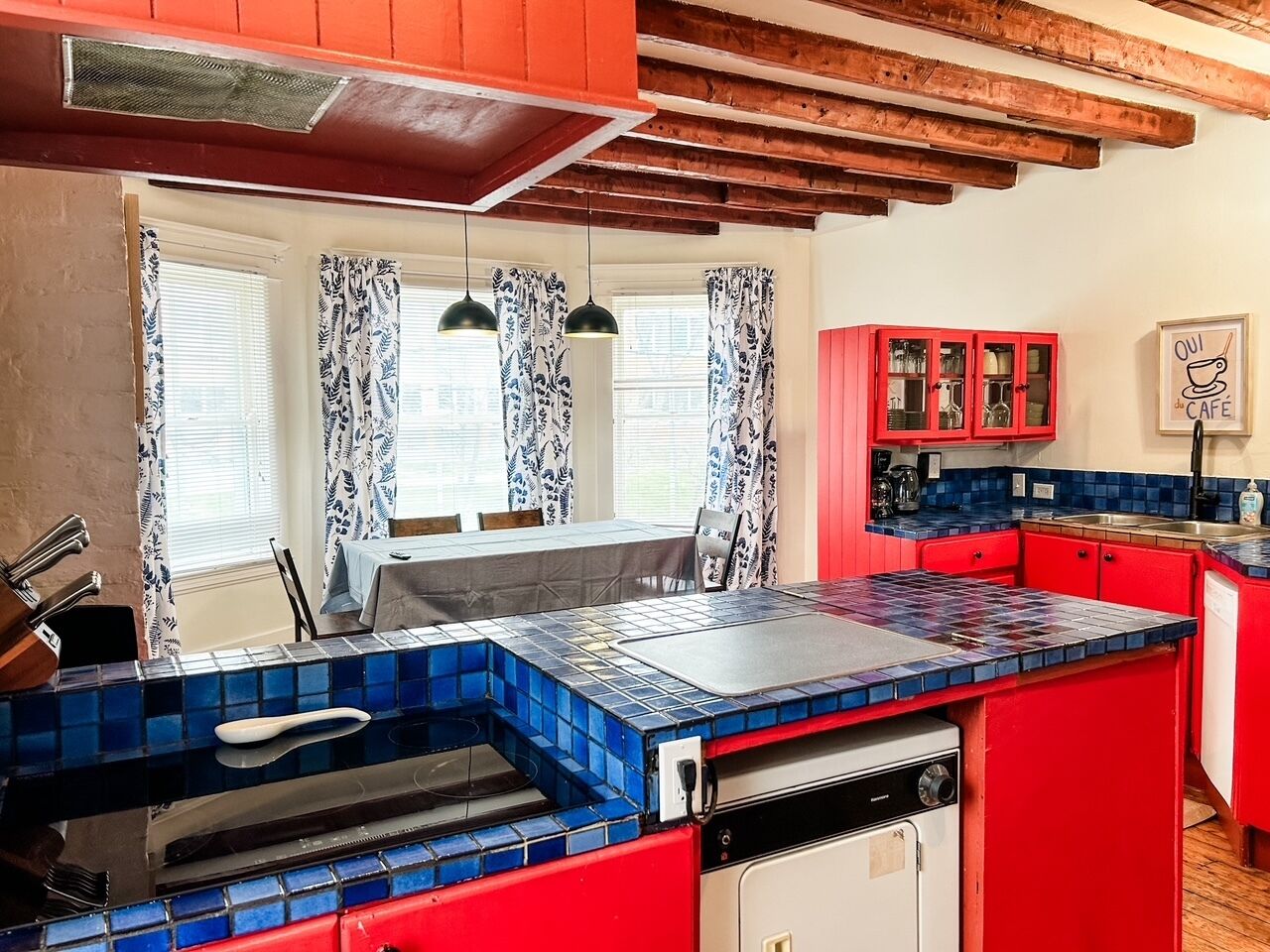 Une cuisine avec des meubles de rangement rouges, des plans de travail recouverts de carreaux bleus et une fenêtre ornée de rideaux à motifs.