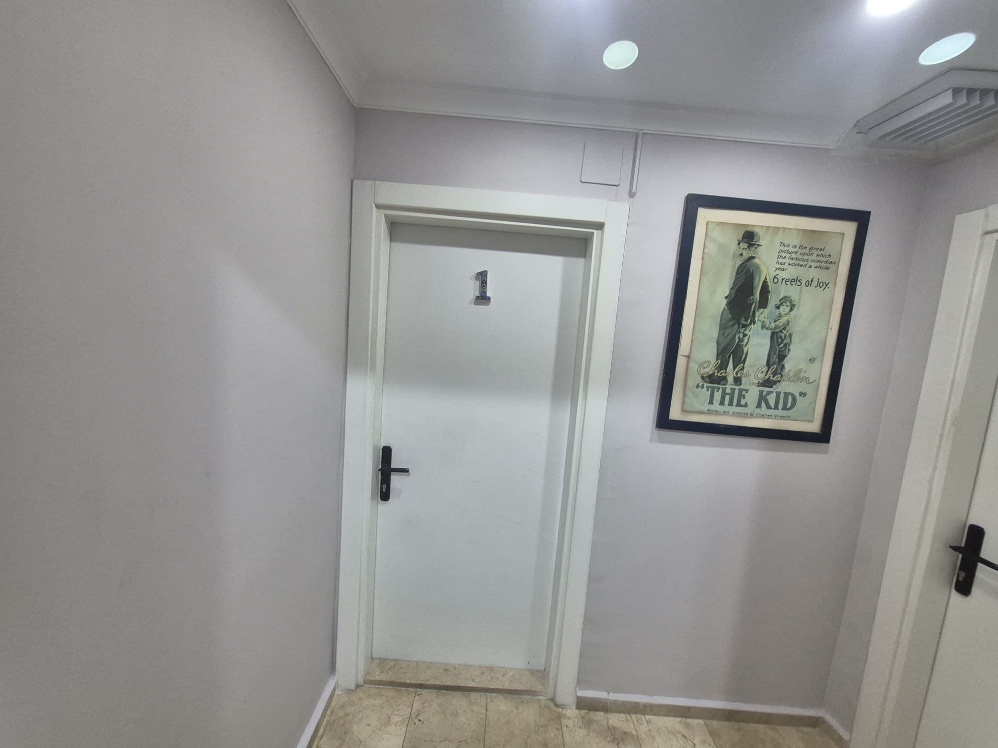 A hallway with a white door marked with the number 1, a framed poster of 'The Kid' on the wall, and a ceiling with recessed lighting.