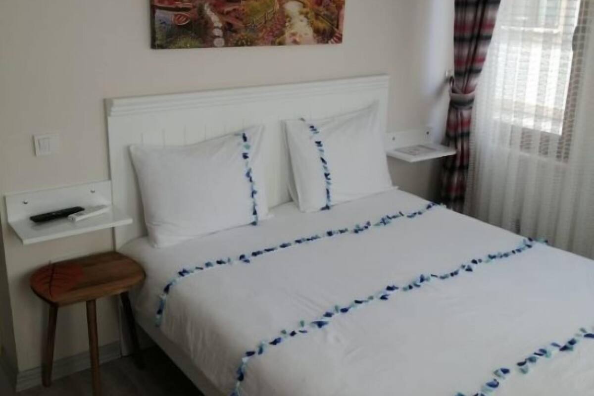 A hotel room with a bed, a painting on the wall, an air conditioner, a remote control, and a window with curtains.