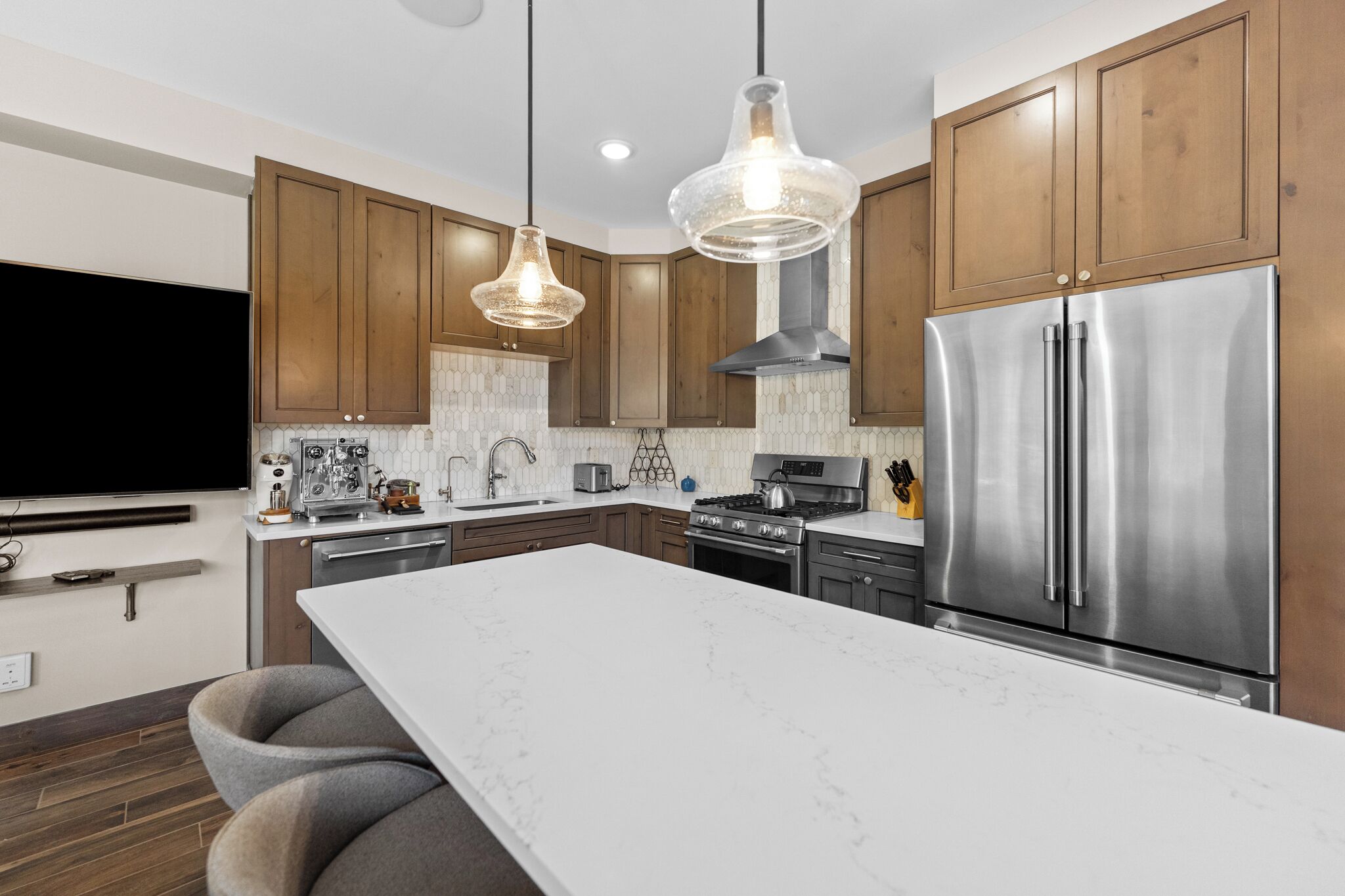 A modern kitchen with wooden cabinets, a stainless steel refrigerator, a large island, pendant lights, and a flat-screen TV.