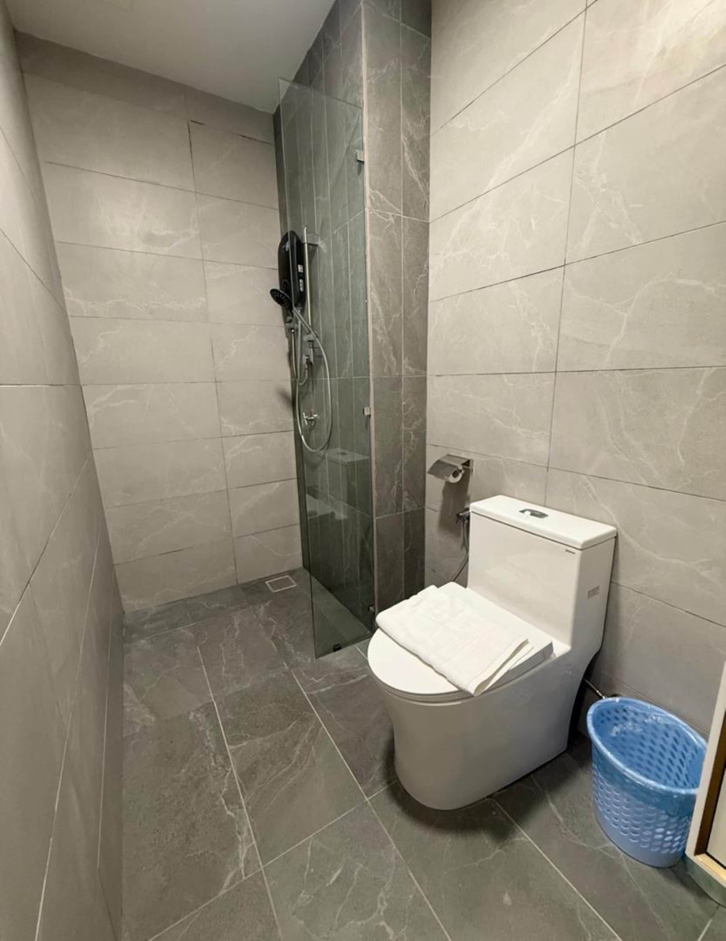 Un baño pequeño con ducha envuelta en vidrio, inodoro y un cubo de basura azul.