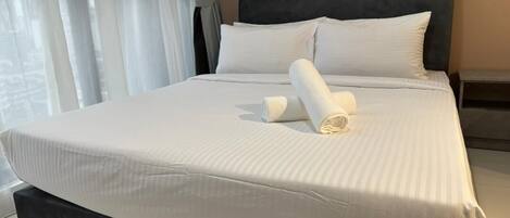 A neatly made bed with white linens and a rolled towel, a dark headboard, and a small bedside table.