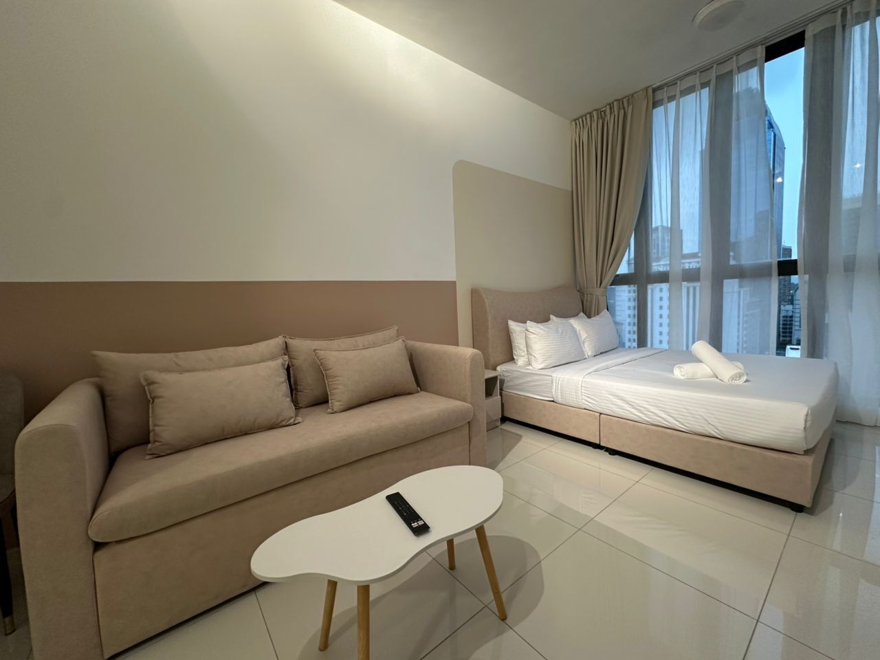 Une chambre d’hôtel moderne avec un canapé beige, une table basse blanche et un lit recouvert de draps blancs.