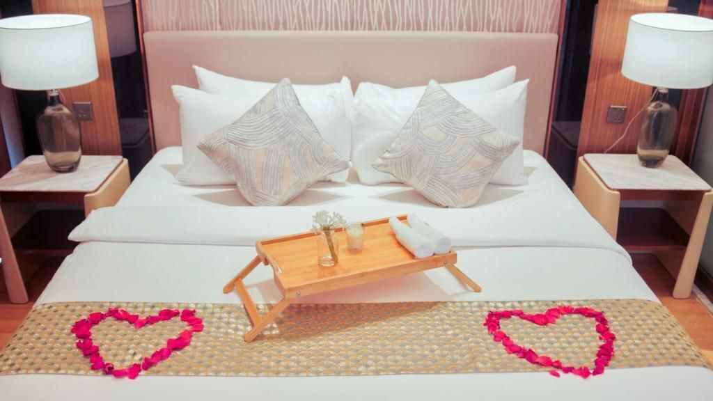 A neatly made bed with a tray of glasses and a bottle, two heart-shaped decorations on the bed, and a wooden headboard.