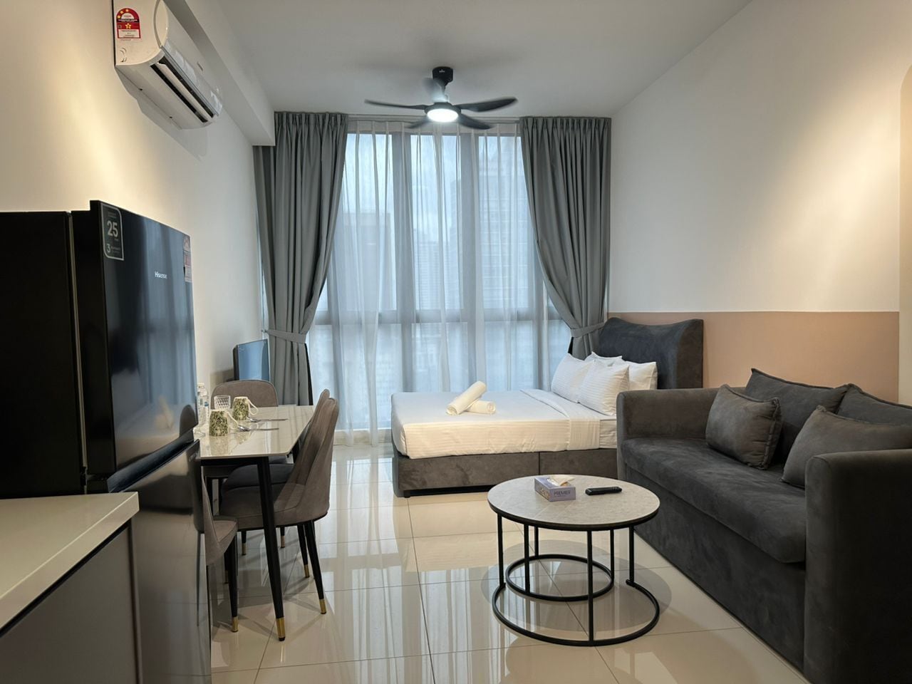 A modern hotel room with a bed, a sofa, a dining table with chairs, a TV, and an air conditioning unit.