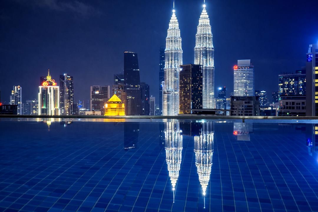 Ein nächtliches Stadtbild mit den Petronas Towers, die hell beleuchtet gegen einen dunklen Himmel hervorstechen und sich auf einer ruhigen Wasseroberfläche spiegeln.