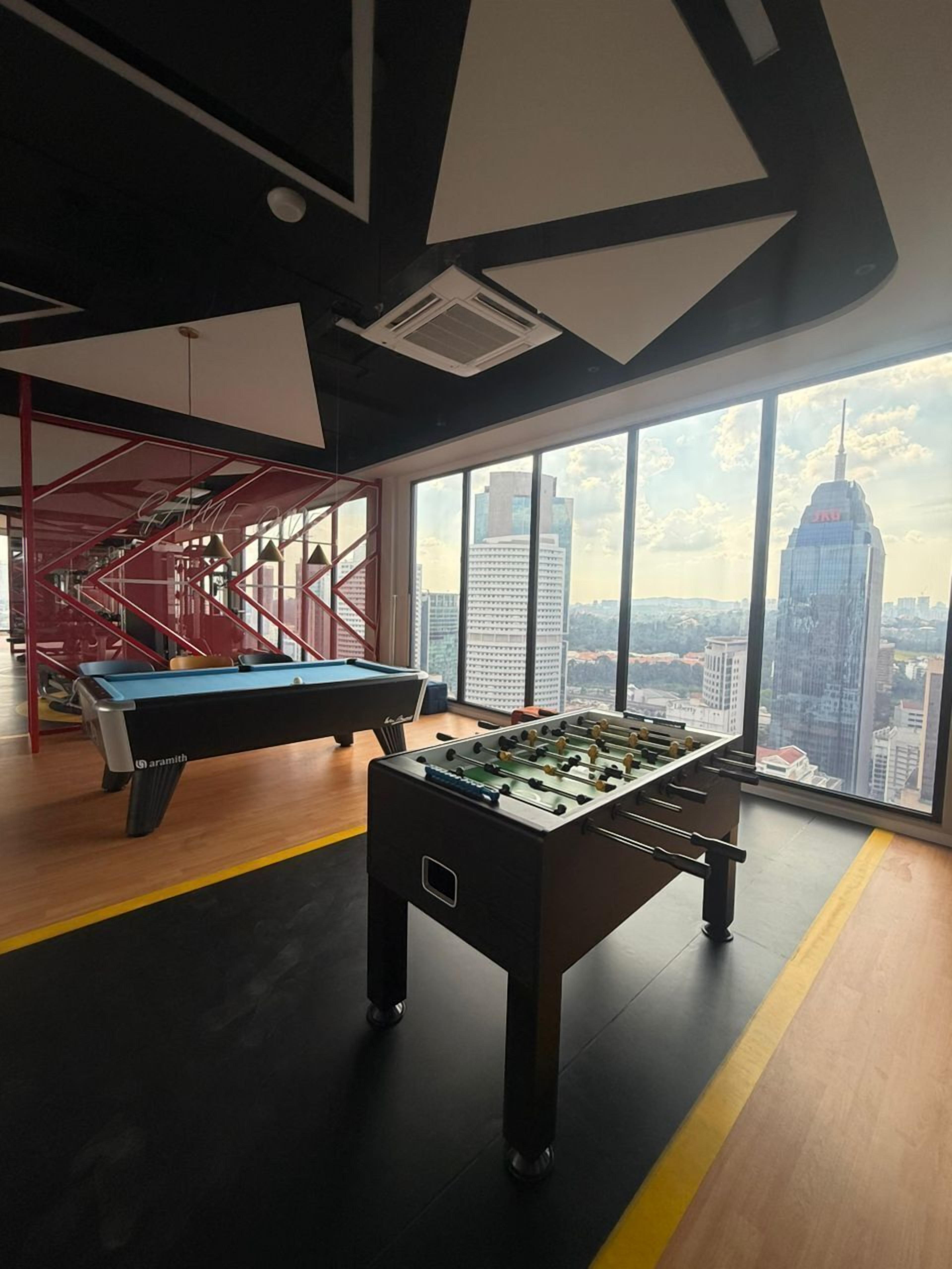 A room with a foosball table, a pool table, and a large window offering a city view.