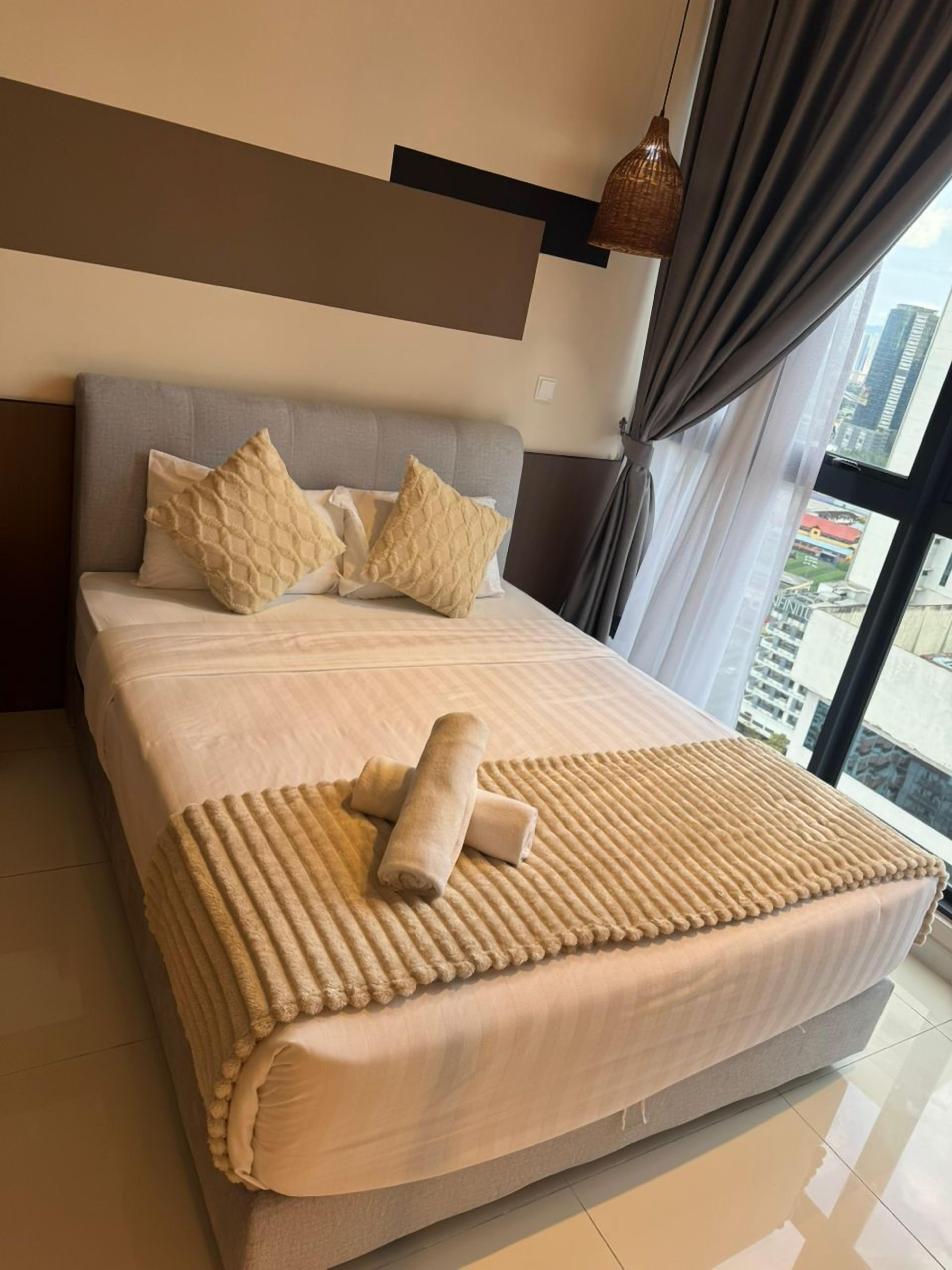 A modern hotel room with a large bed, a headboard, and a view of the city through large windows.