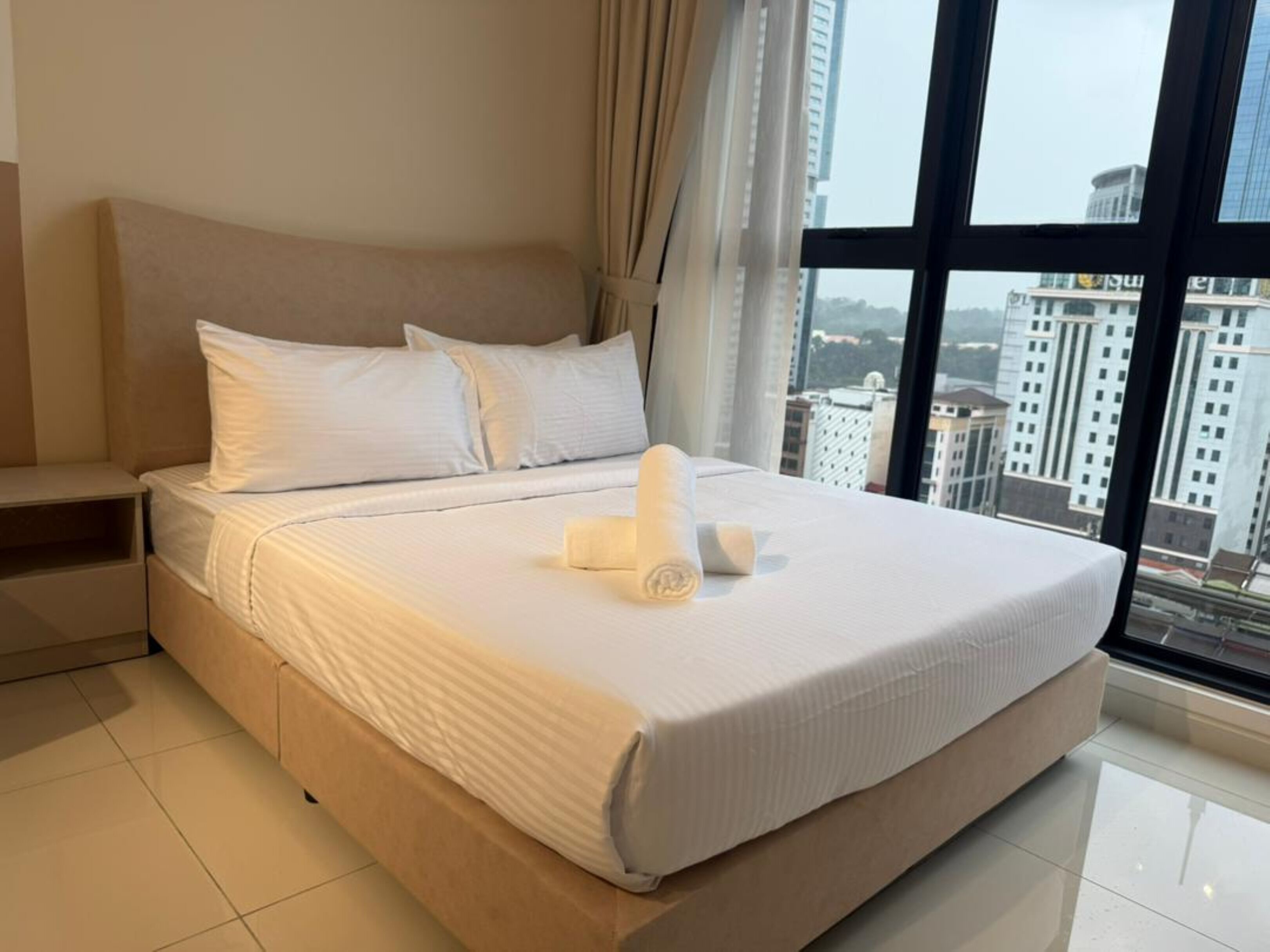 Une chambre d’hôtel moderne avec un grand lit, une table de chevet et une vue sur la ville grâce à de grandes fenêtres.