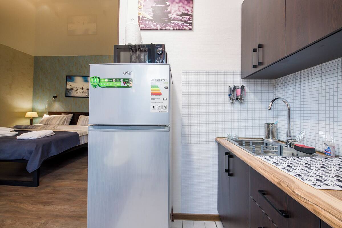 Een compacte hotelkamer met kitchenette, een bed met blauwe sprei, een klein nachtkastje met lamp en een wanddecoratie.