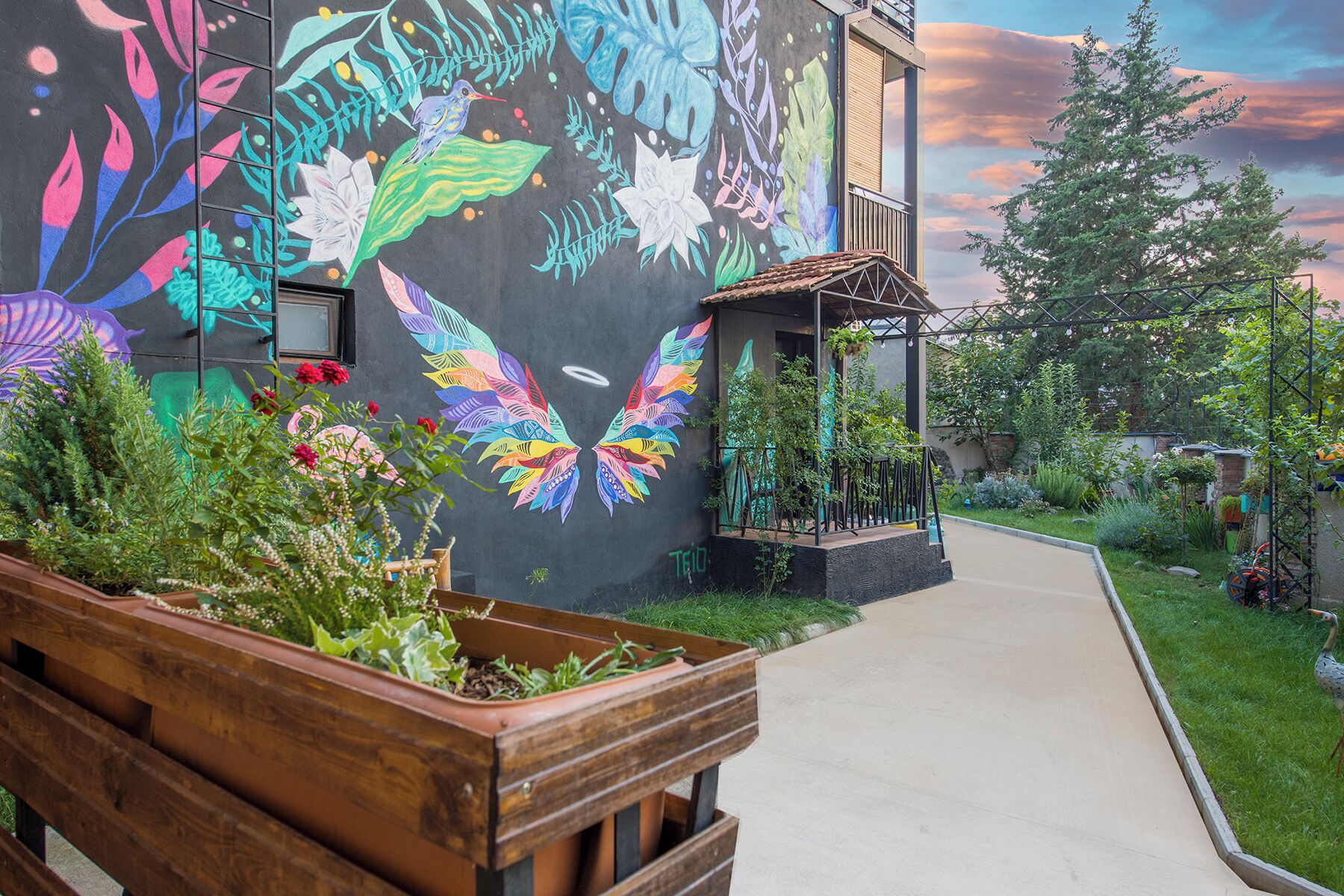 Un edificio con un mural colorido, un banco de madera y un jardín con diversas plantas.