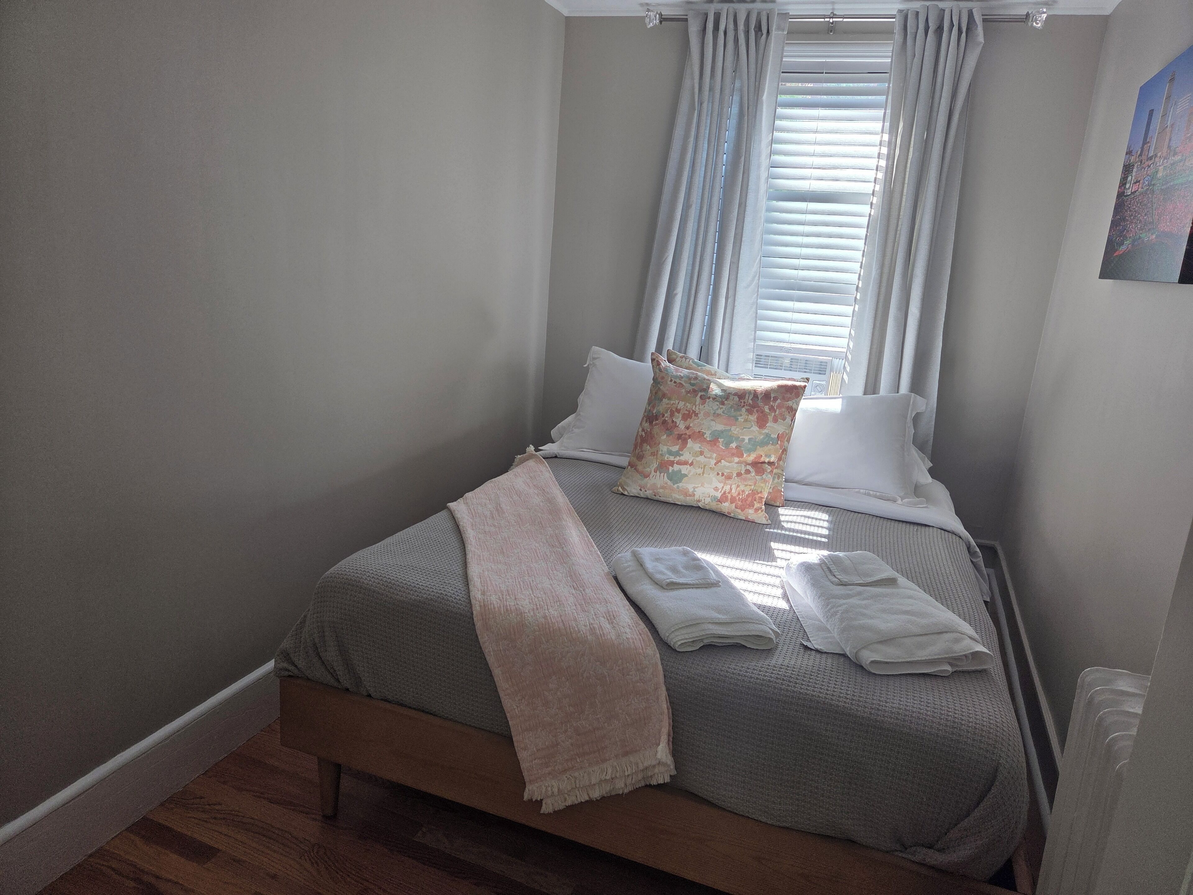 A neatly made bed with a gray blanket and pillows, a beige throw blanket, and neatly folded towels on the side.