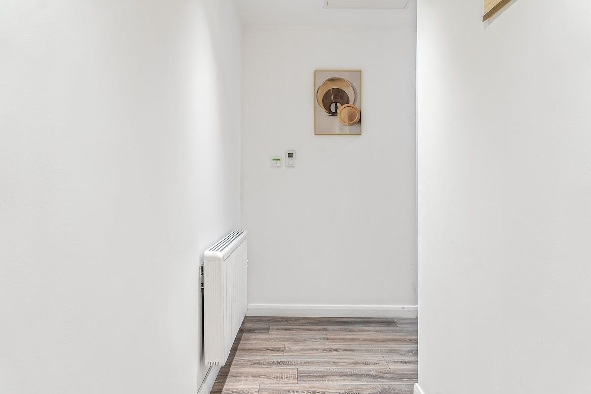 Un couloir étroit avec un radiateur, un tableau encadré accroché au mur et un interrupteur.