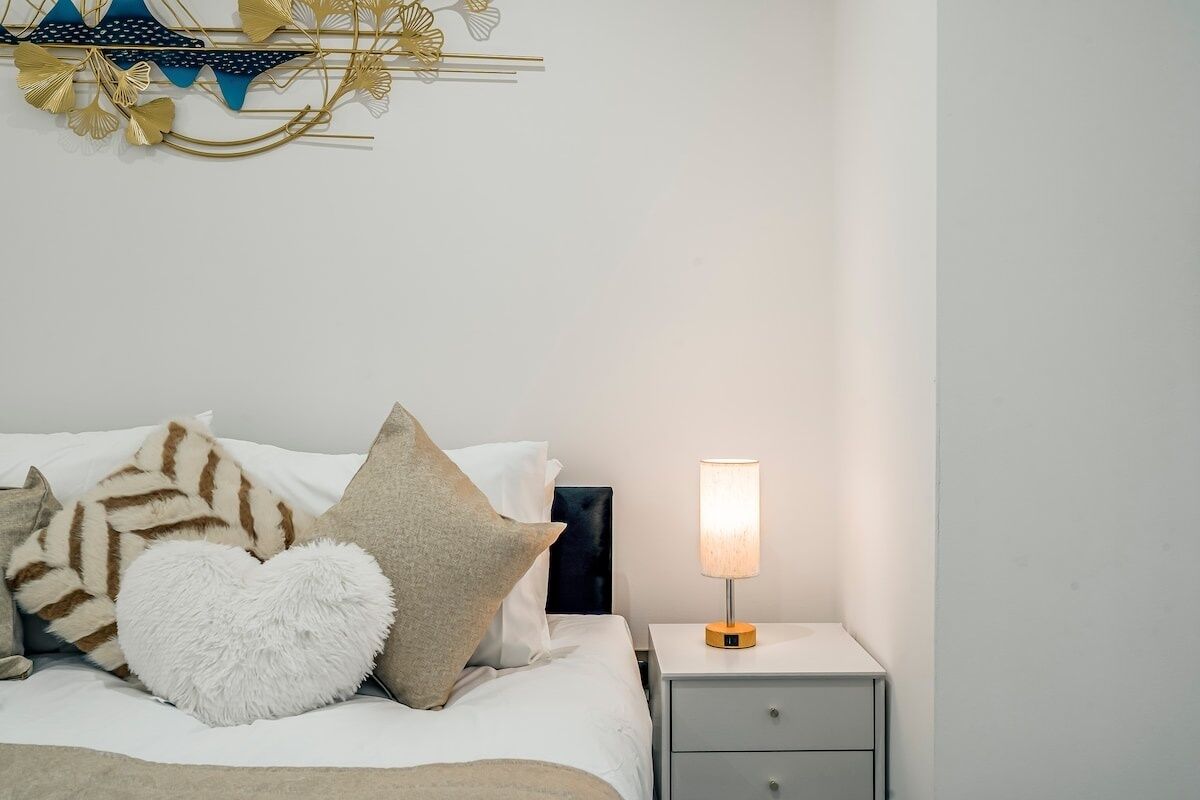A neatly made bed with a white and brown striped blanket, a white fluffy pillow, and a heart-shaped pillow. A bedside lamp with a white shade is on a small bedside table.