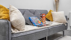A grey sofa with multiple cushions in various colors, a magazine rack with books, and a vase with dried plants. - The Azure Hideaway 2BR 2BA City Centre (Leicester)