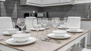 A modern dining area with a rectangular table set for four, white chairs, and a kitchen in the background. - The Azure Hideaway 2BR 2BA City Centre (Leicester)