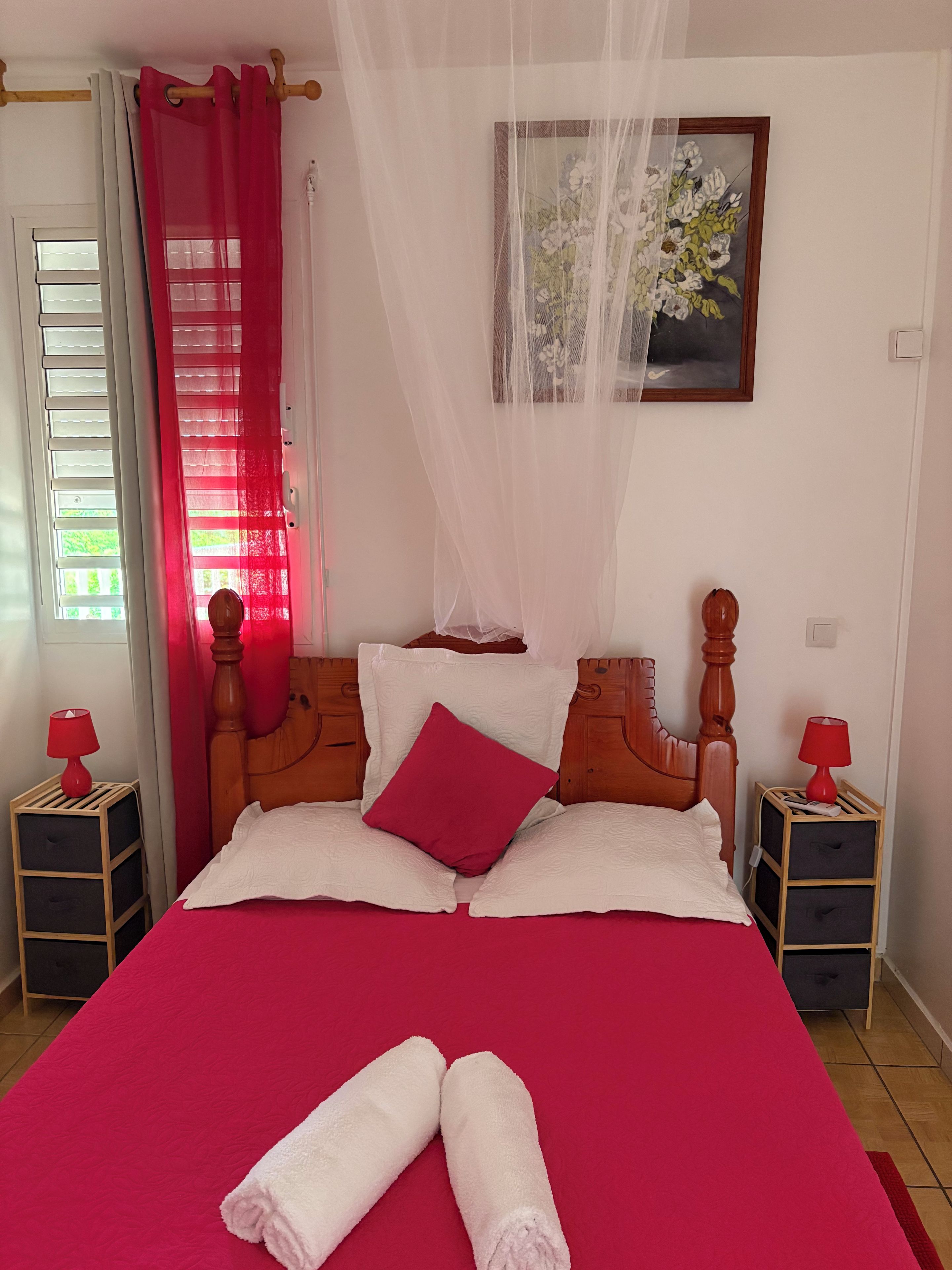 Ein ordentlich bezogenes Bett mit roter Bettdecke und weißen Kissen, ein Holz-Kopfteil, ein Nachttisch mit roter Lampe und ein Fenster mit weißen Fensterläden.