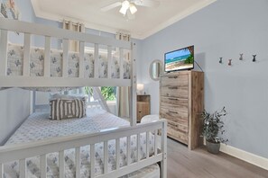 Un lit superposé pour enfant, avec une couverture à motifs, une commode en bois avec des tiroirs, un téléviseur posé dessus et une plante en pot.