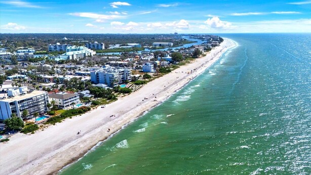An aerial view of a coastal city with a long sandy beach, adjacent buildings, and a clear blue sky.