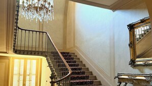Uma escadaria imponente com grades ornamentadas, um candelabro e tapetes estampados.