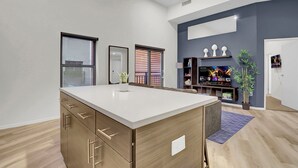 A modern kitchen with a central island, wooden cabinets, and a built-in oven. There is a dining area with a table and chairs, a large window with blinds, and a decorative shelf with plants.