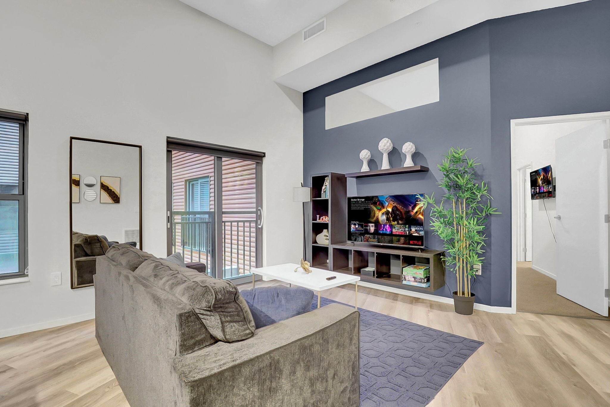 Una sala de estar moderna con un sofá gris, una mesa de centro blanca y un televisor de pantalla plana sobre un mueble. Hay un ventanal con persianas, una planta en maceta y una estantería con objetos decorativos.