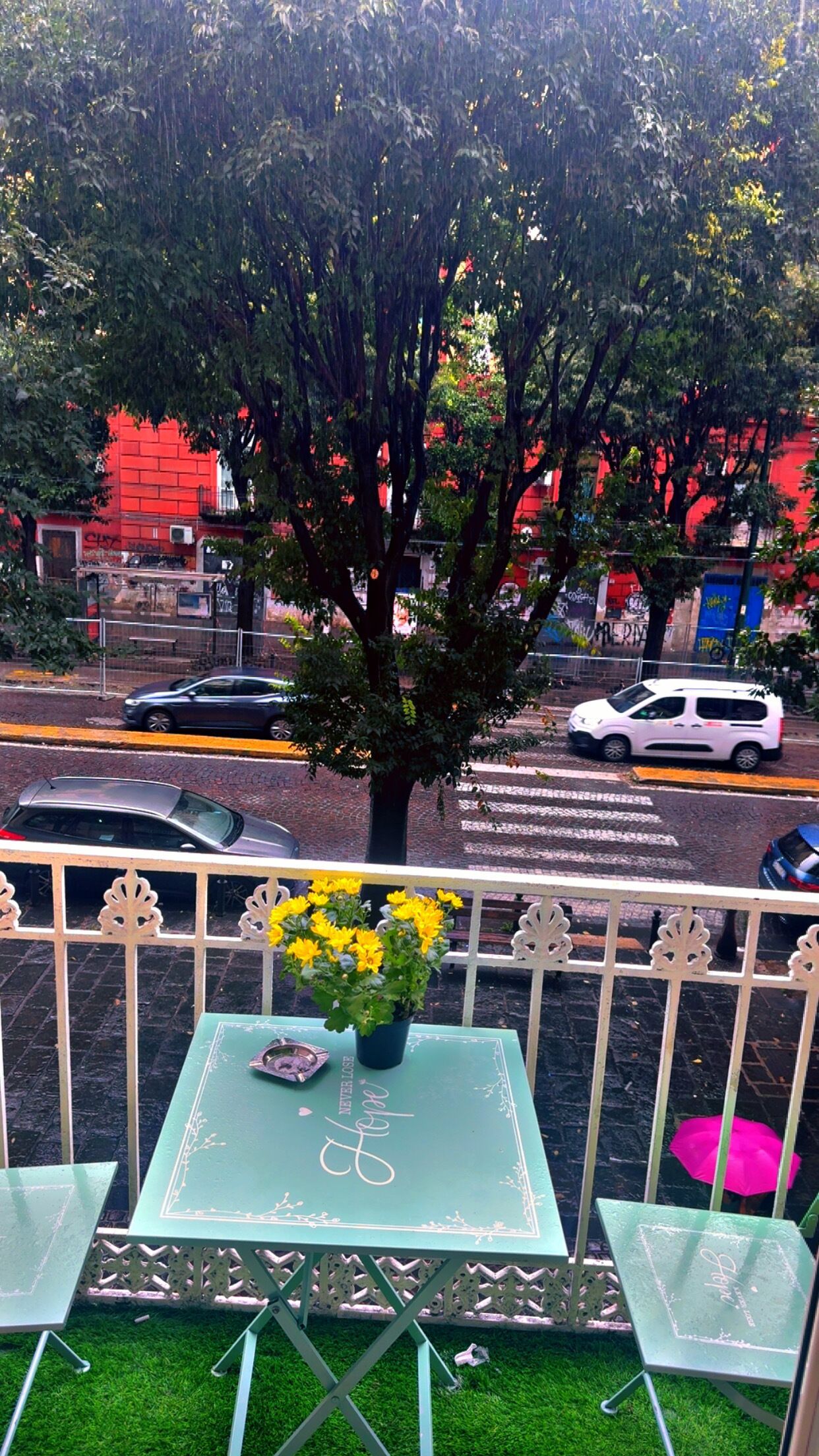 Een balkon met een tafel en stoelen, een bloempot met gele bloemen, en uitzicht op een straat met auto's en gebouwen.