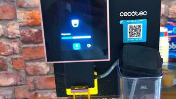 Eine Kaffeemaschine mit digitalem Display, einem QR-Code für den App-Zugriff und einer Kaffeetasse.