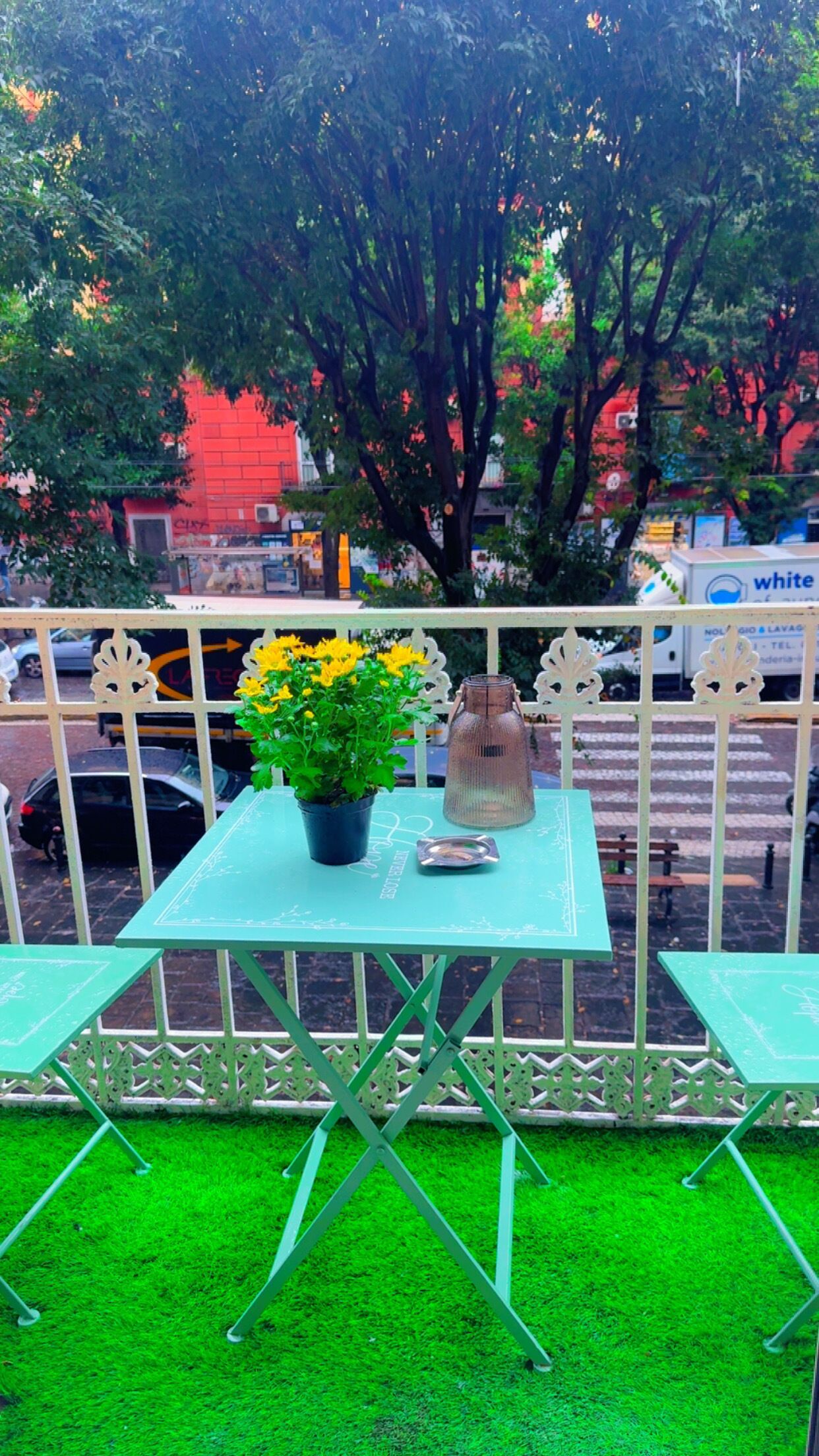 A green outdoor seating area with a table and chairs, a potted plant with yellow flowers, and a decorative vase.