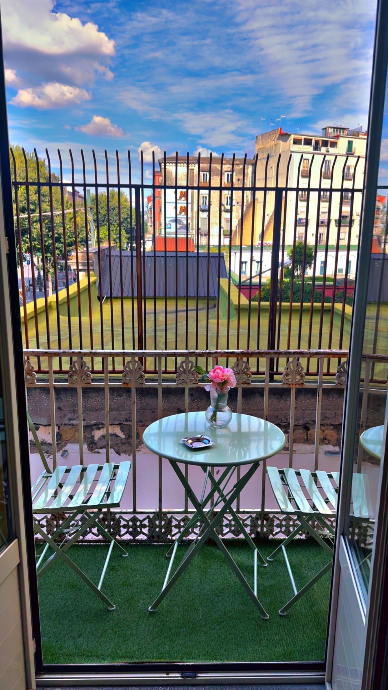 Ein Balkon mit Glastisch und zwei Stühlen, einer Blumenvase sowie Blick auf Gebäude hinter einem Metallzaun.