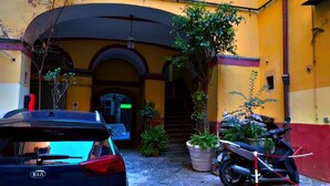 A courtyard with a blue car parked in front of a building with a yellow facade, a motorcycle, and potted plants. - هوستيل دولشي دي مونيكا نابوليتنا (Napoli)