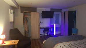 Hotellihuone, jossa on sänky, työpöytä tuolilla, vaatekaappi, seinälle asennettu televisio ja yöpöytä lampulla.