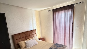 A bedroom with a bed, a headboard, a nightstand, a window with curtains, and a ceiling light. - Your Holiday Home in Agadir (Agadir)