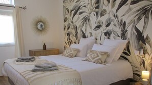 A neatly made bed with a white bedspread and multiple pillows, a bedside table with a vase, and a wall with a floral wallpaper pattern.
