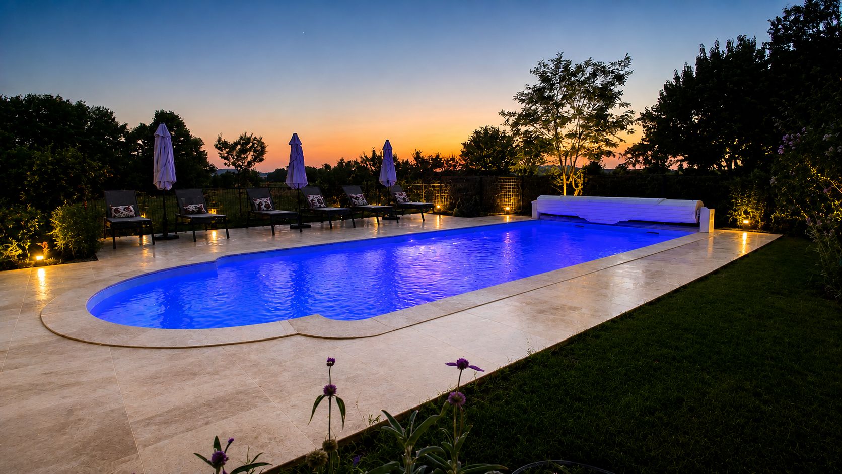 Une piscine à bord incurvé, entourée de chaises longues et de parasols, et une pelouse agrémentée de fleurs.