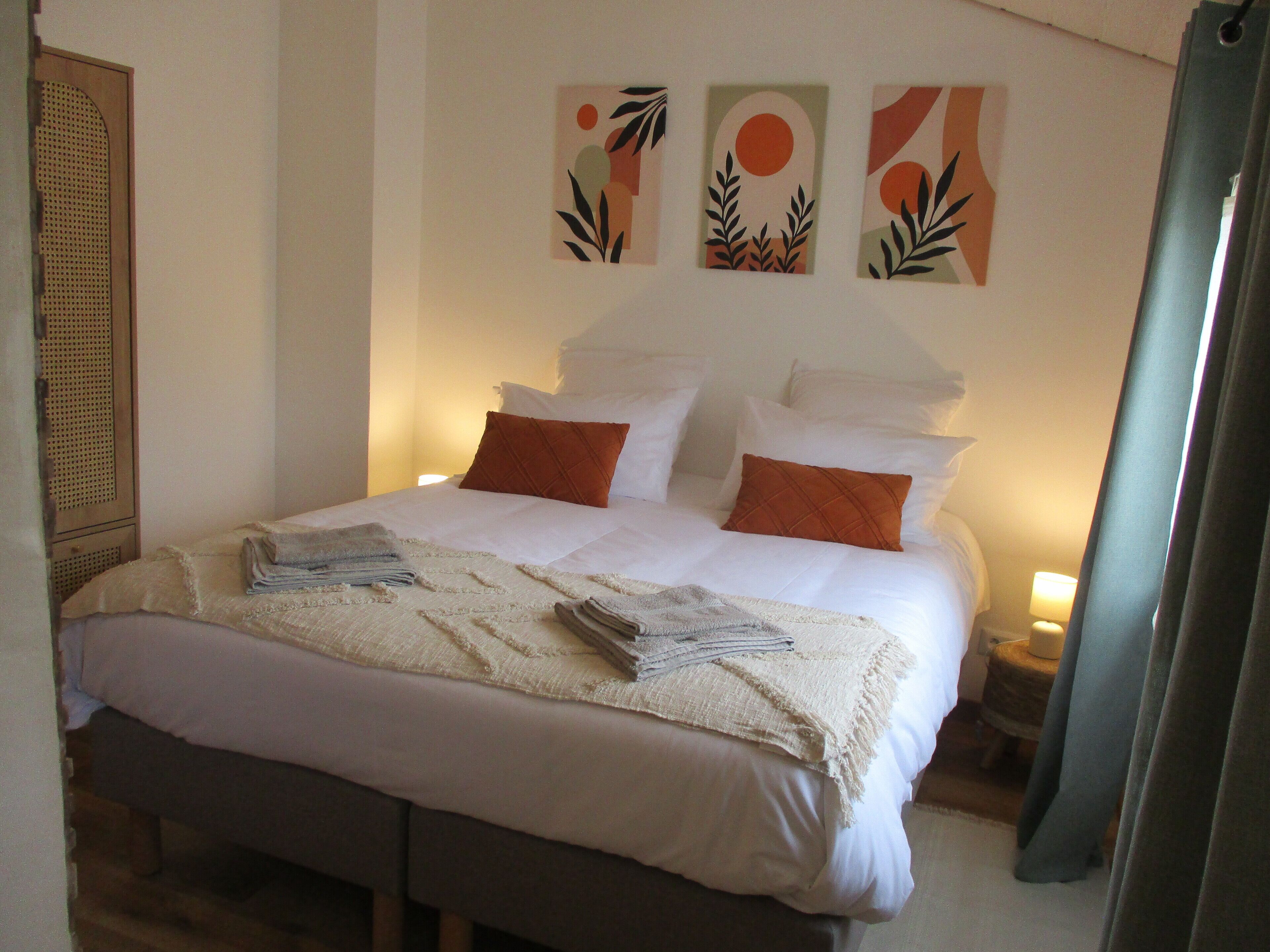 Una cama bien tendida con sábanas blancas y dos almohadas naranjas, una mesita de noche con una lámpara, y tres obras de arte abstracto en la pared.