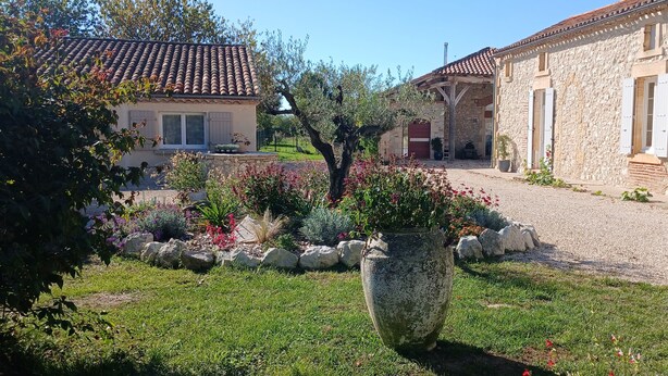A stone building with a tiled roof, a gravel driveway, and a garden with various plants and flowers.