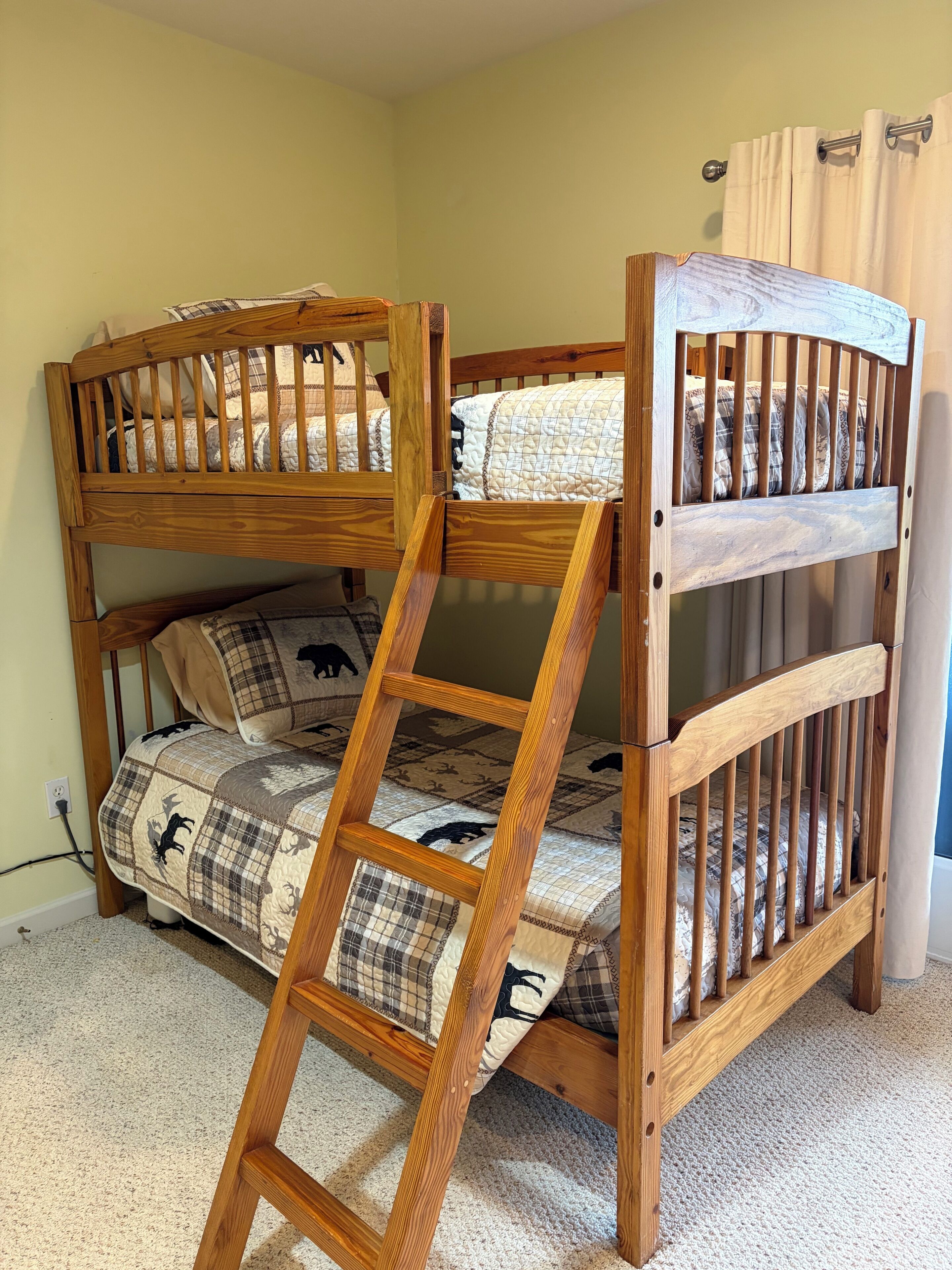 A bunk bed with a wooden ladder, a quilted mattress, and a plain wall with a curtain.