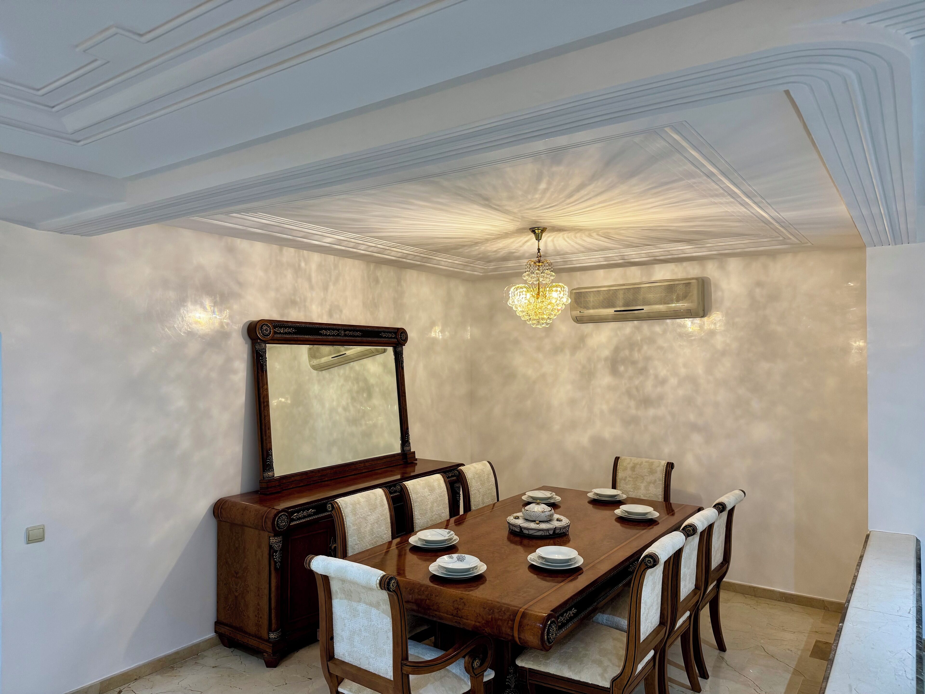 A dining room with a wooden table, chairs, a chandelier, and a large mirror.
