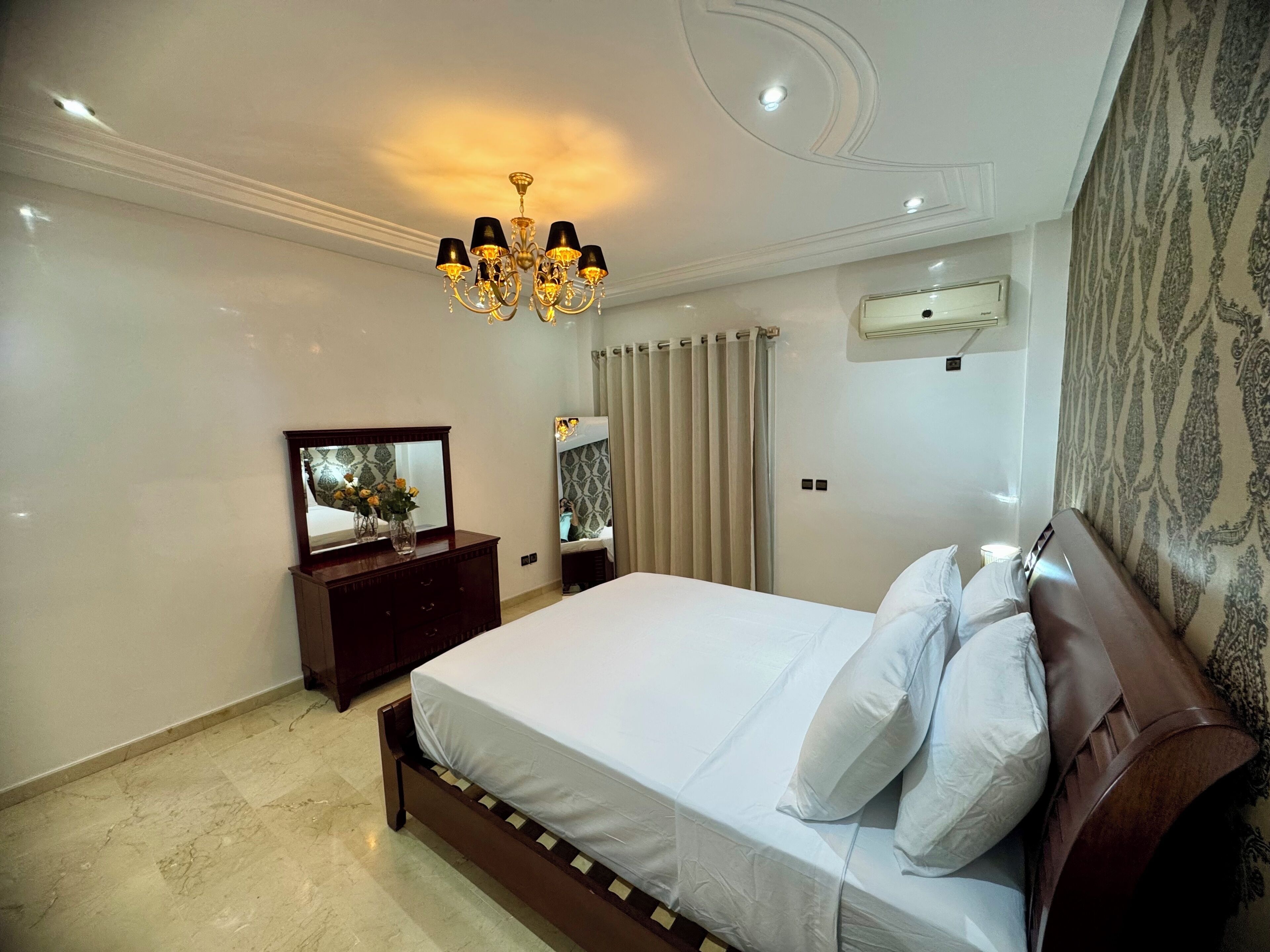 A bedroom with a large bed, a dresser, a mirror, a chandelier, and a window with curtains.