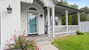 Uma casa com fachada branca, uma porta da frente, varanda com grades brancas e um pequeno jardim com flores cor-de-rosa.
