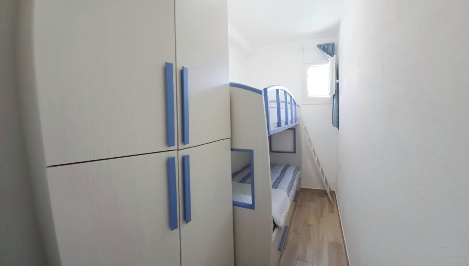 A narrow room with a bunk bed, white walls, and blue handles on the wardrobe.