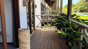 Una terraza de madera con barandilla, un poste de madera y vista a un edificio con un corredor cubierto.