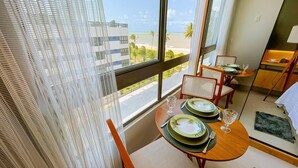 A hotel room with a balcony overlooking a beach and palm trees. There is a dining area with a table set for two, including plates, glasses, and cutlery. - Setai Aquamaris Vista Mar Lateral (João Pessoa)