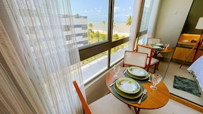 A hotel room with a balcony overlooking a beach and palm trees. There is a dining area with a table set for two, including plates, glasses, and cutlery.