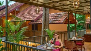 A dining area with a table set for two, surrounded by wicker chairs, with a view of traditional buildings in the background. - MALABAR KOVILAKAM (Tirurangadi)