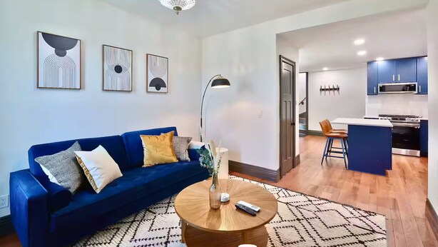 A modern living room with a blue sofa, a round wooden coffee table, and abstract wall art. A kitchen with blue cabinets and a dining area with a blue island and chairs are visible in the background.