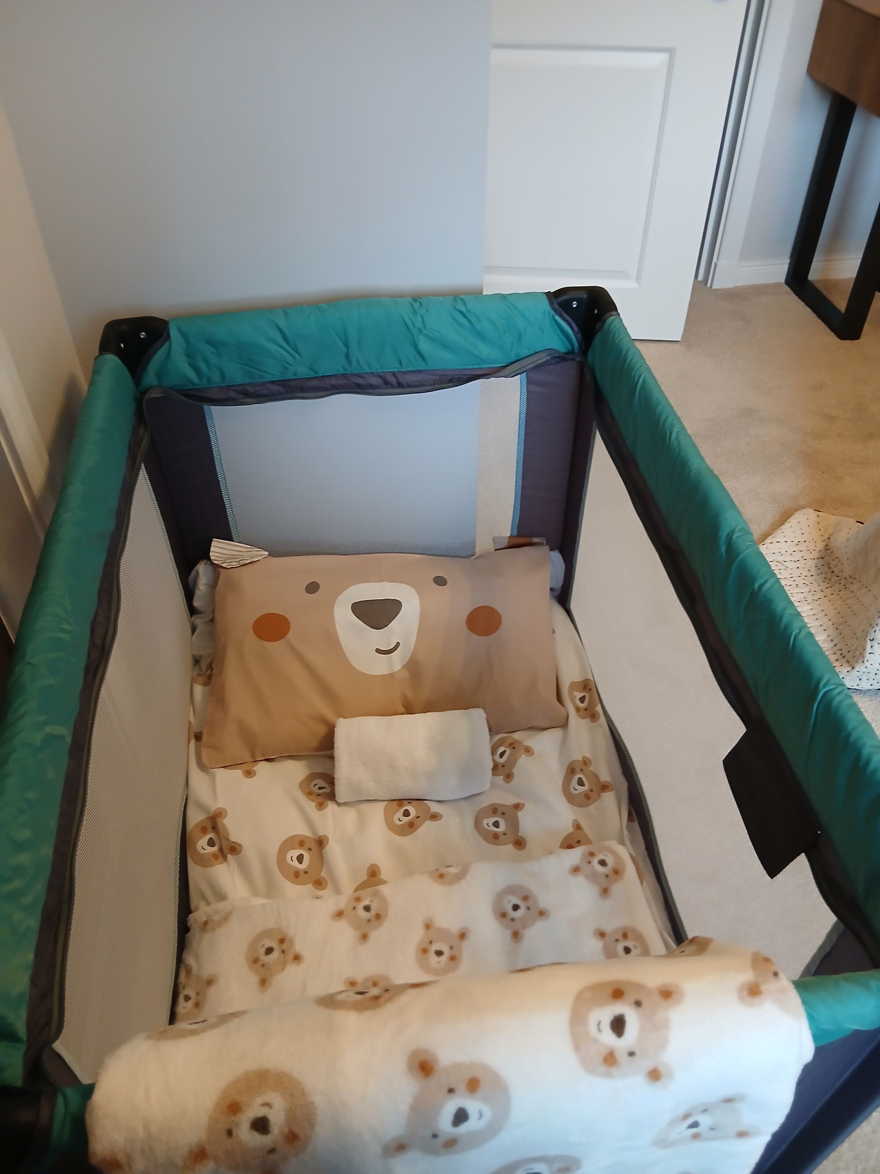 Ein Babybett mit tealfarbener Rahmung, Bettwäsche mit Beige- und Brauntönen im Teddybären-Design und eine weiße Tür im Hintergrund.